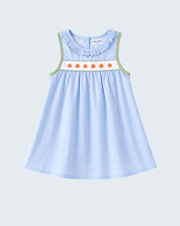 A light blue sleeveless baby dress with a ruffled neckline, featuring a white smocked band with orange pumpkin motifs and sage green trim along the armholes, shown against a light gray background.