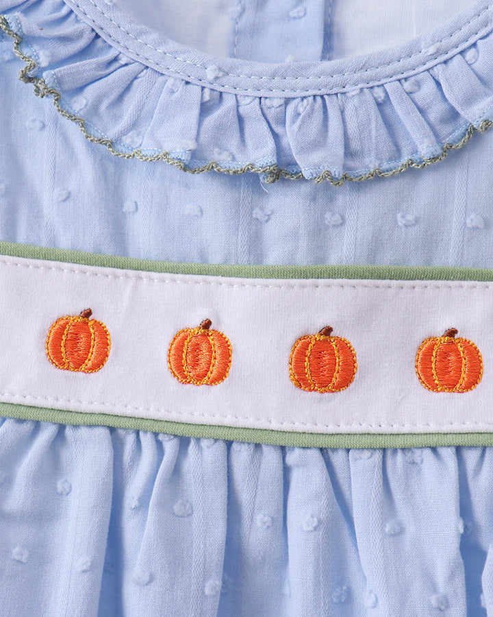 Close-up of a light blue baby dress with a ruffled neckline, a white horizontal band featuring four embroidered orange pumpkins, and a thin light green trim.