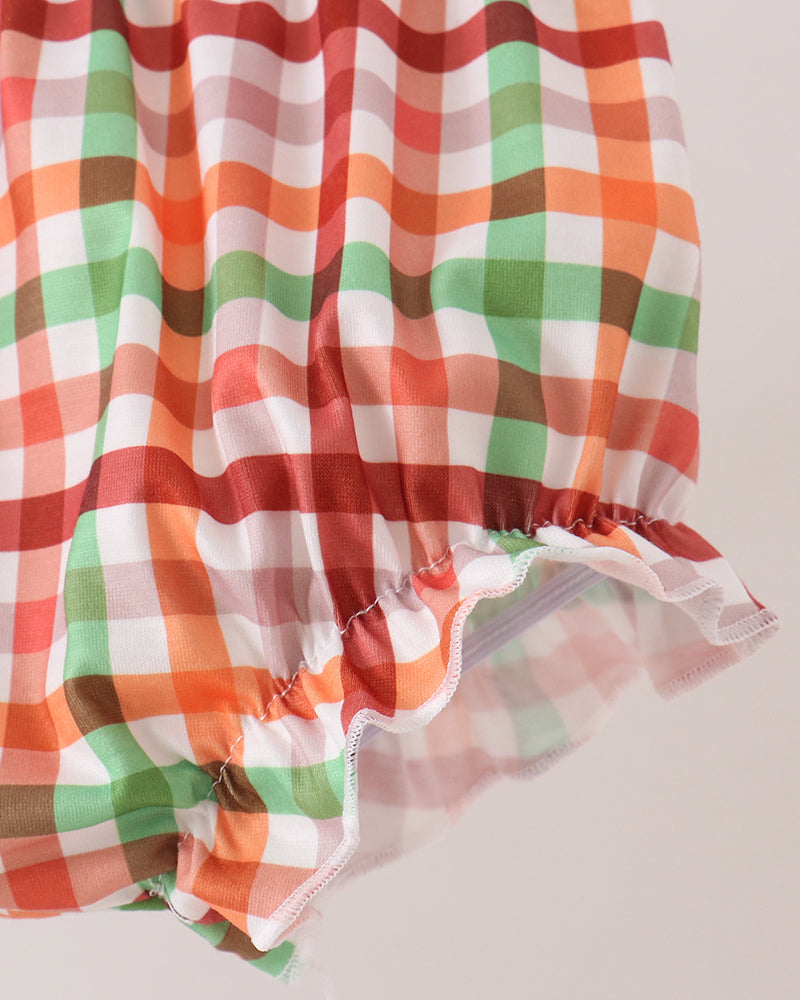 A close-up of a sheer, ruffled fabric with a multicolor plaid pattern in orange, green, brown, and white, set against a solid light beige background.