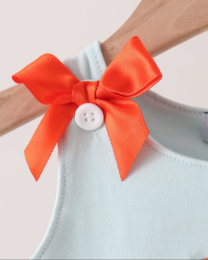 A close-up of a light blue knit top with a bright orange satin bow and a white button accent, hanging on a wooden hanger against a warm beige background.