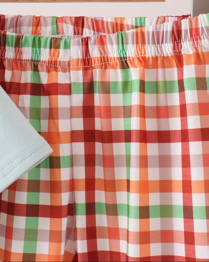 A close-up of plaid pants in orange, green, red, and white, hanging on a wooden hanger with a light blue garment partially visible in the foreground.