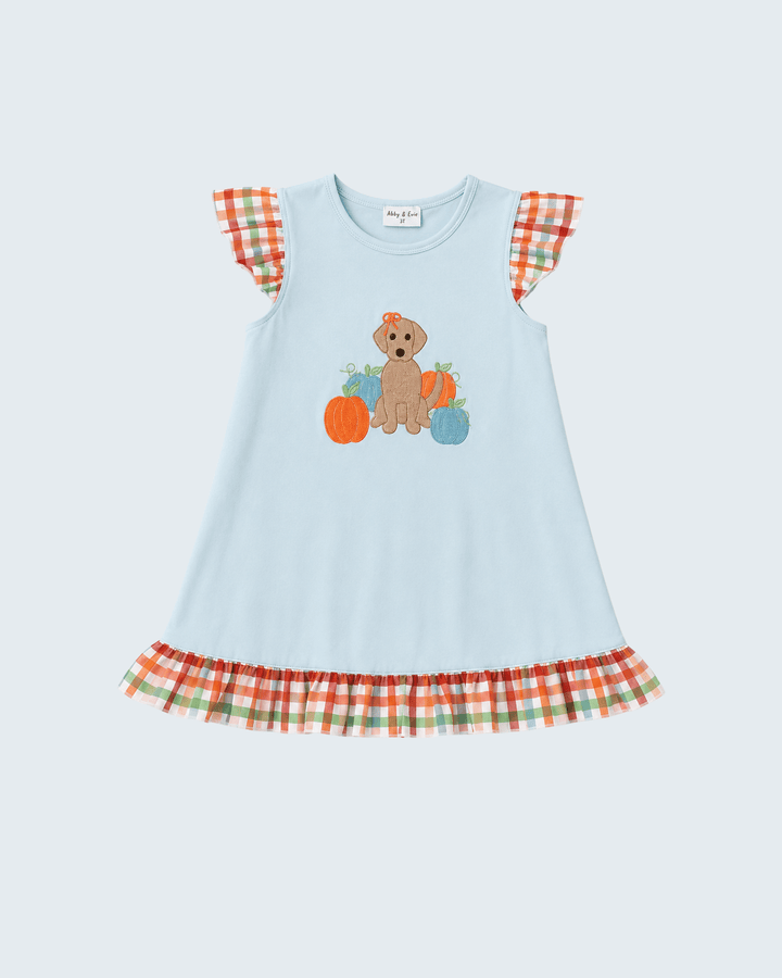 A light blue sleeveless dress with a fall-themed graphic of a puppy and pumpkins on the front, featuring orange, green, and white plaid ruffle details on the shoulders and hem.