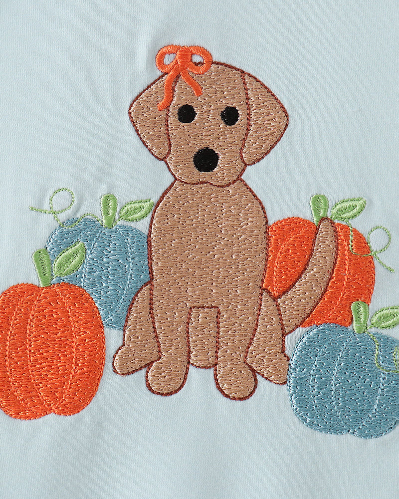 Embroidered autumn dog design featuring a brown Labrador retriever with an orange bow, surrounded by orange pumpkins and blue gourds on a white fabric background.
