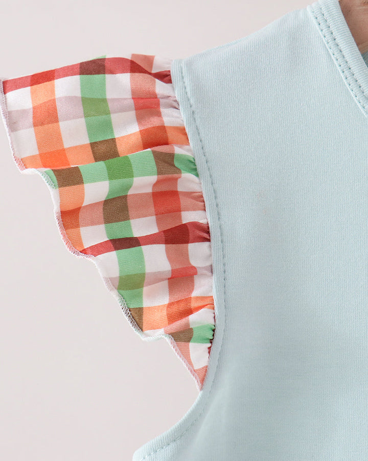 Close-up of a light blue sleeveless top with a ruffled shoulder detail featuring a red, green, orange, and white gingham pattern.