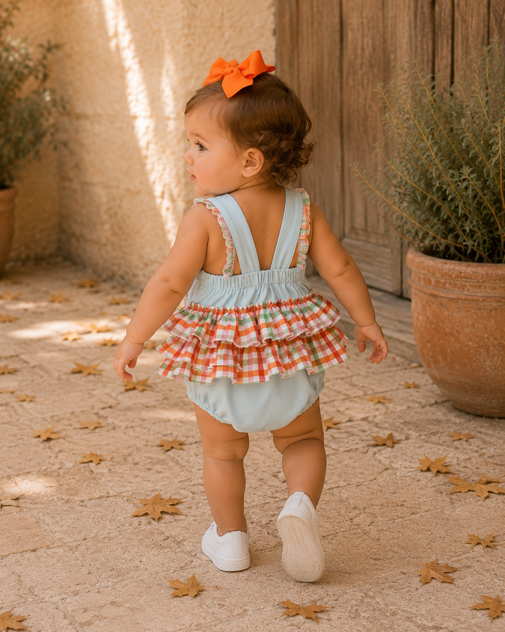A toddler girl walking away in a light blue ruffled romper with a red and white gingham tiered skirt, wearing a bright orange bow in her hair, on a stone patio with potted plants.