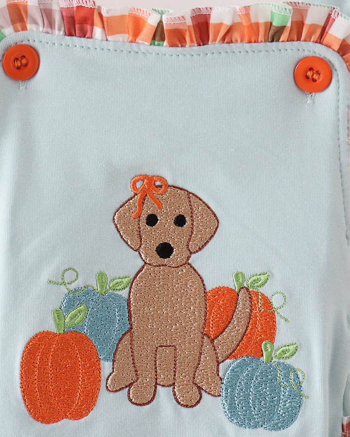 A close-up of a light blue fabric item, likely a bag or pouch, featuring an embroidered autumn design of a brown puppy sitting among orange and blue pumpkins. The top edge has orange and white striped ruffles with two orange buttons.