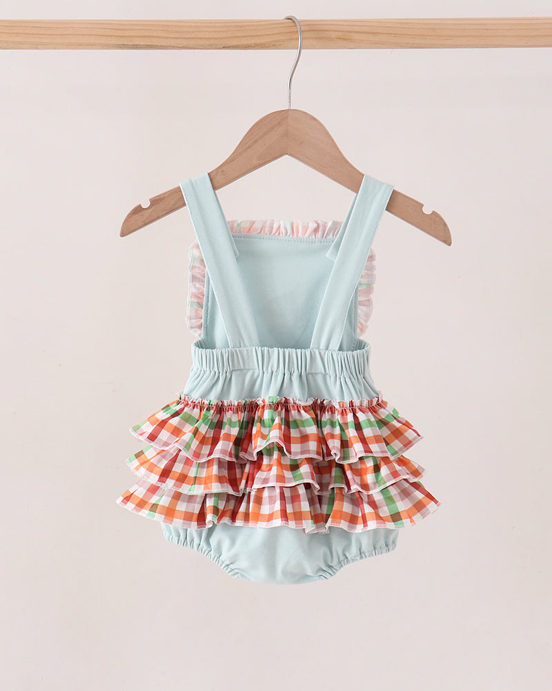 A baby romper with a light blue mint body and a tiered ruffle skirt in a red, green, and white gingham pattern, hanging on a wooden hanger against a light beige background.
