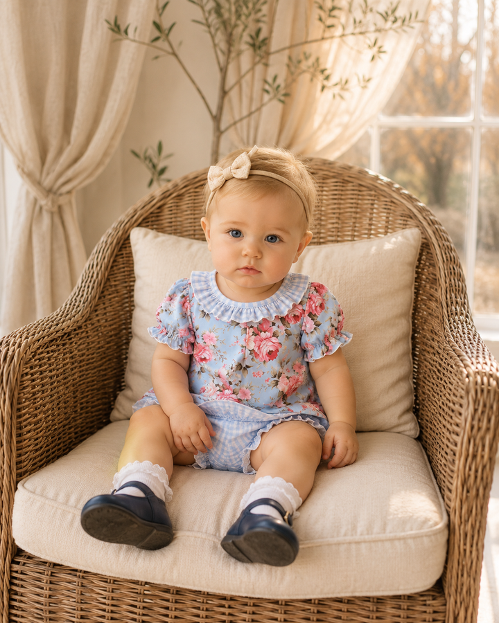 A baby girl wearing a light blue floral romper and matching checkered shorts sits on a wicker armchair with cream cushions in a sunlit room.