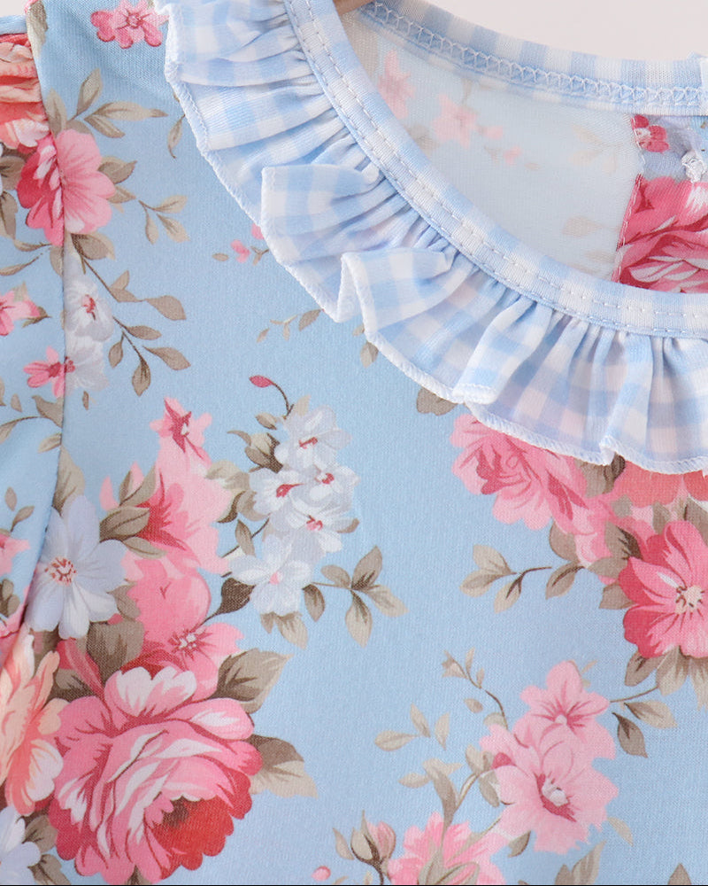 Close-up of a light blue floral top with pink and white flowers, featuring a light blue and white gingham ruffled collar, hanging on a wooden hanger.