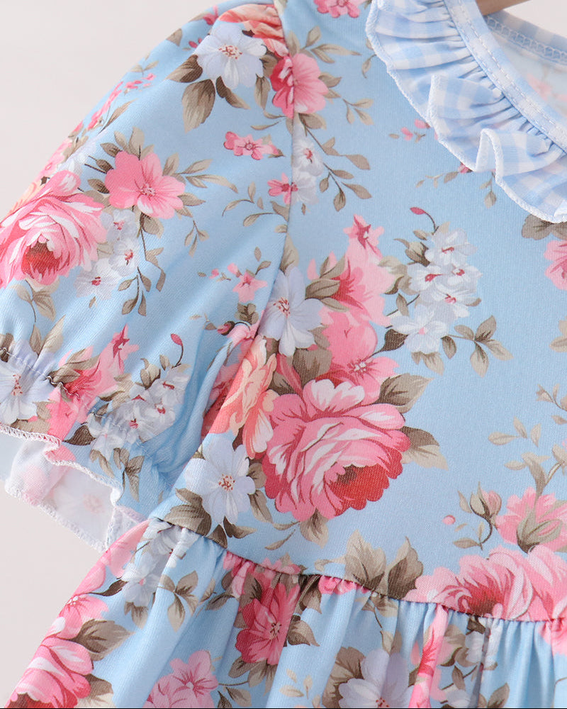 A close-up of a light blue floral dress featuring pink and white roses with green leaves, a ruffled light blue collar, and a tiered skirt, shown on a wooden hanger.