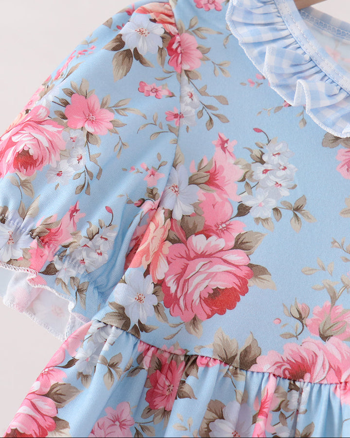 A close-up of a light blue floral dress featuring pink and white roses with green leaves, a ruffled light blue collar, and a tiered skirt, shown on a wooden hanger.
