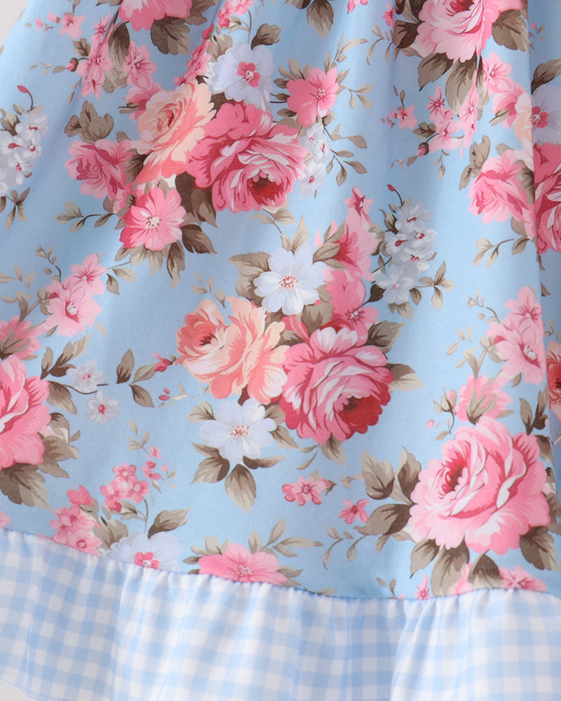 A close-up of a light blue floral dress featuring pink and white roses with green leaves, finished with a light blue and white gingham ruffled hem.