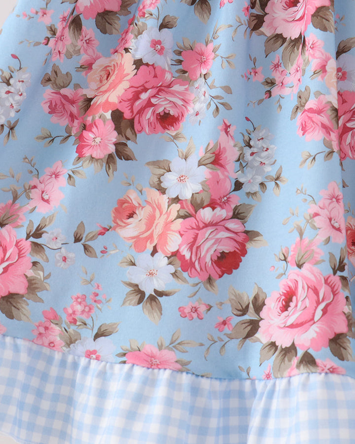 A close-up of a light blue floral dress featuring pink and white roses with green leaves, finished with a light blue and white gingham ruffled hem.