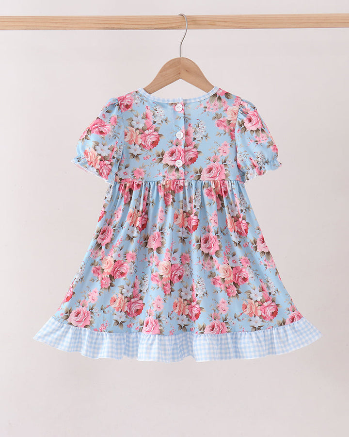A light blue short-sleeve dress with a pink rose floral print, hanging on a wooden hanger against a plain beige wall. The dress features a smocked bodice, buttoned back, and a light blue and white gingham checkered ruffle hem.