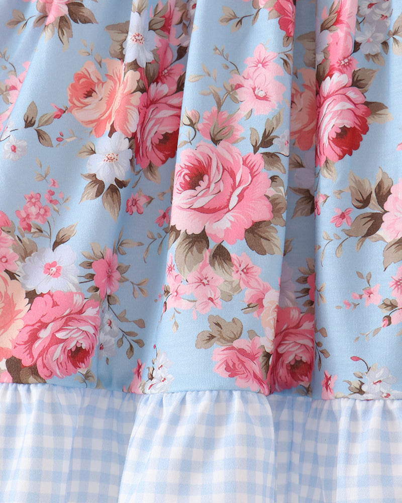 Close-up of a light blue floral dress with pink and white roses, green leaves, and a light blue gingham ruffled hem.