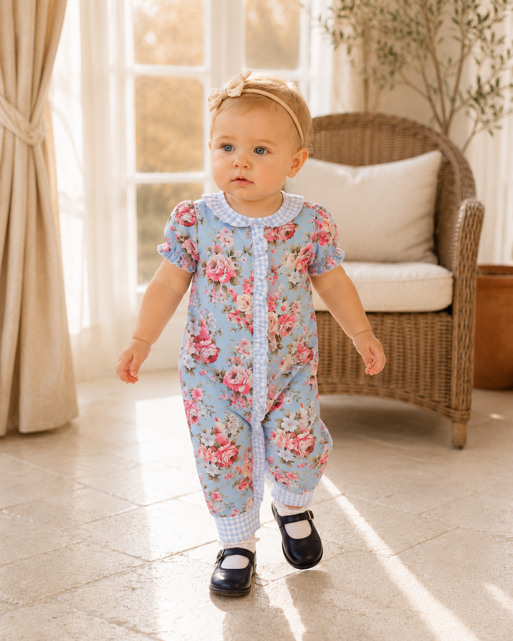 A toddler girl wearing a light blue short-sleeve floral jumpsuit with a white gingham collar and cuffs, walking indoors on a tiled floor near a wicker chair and window.