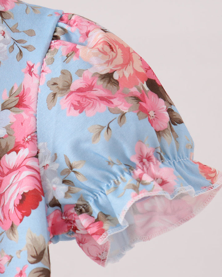 Close-up of a light blue floral blouse with pink and red roses, brown leaves, and ruffled short sleeves on a plain light background.
