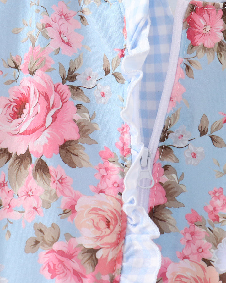 Close-up of a baby garment featuring a light blue background with a pink rose floral print, a white and blue gingham panel, and white ruffle trim along a front zipper.