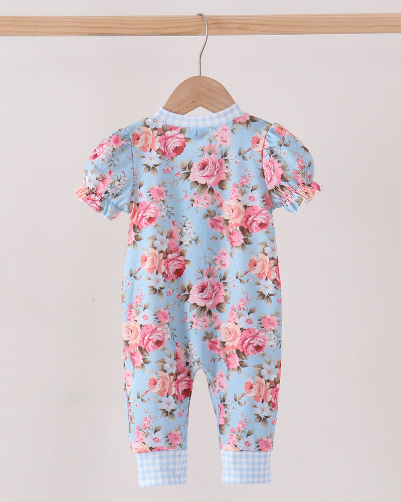 A baby jumpsuit with a light blue background and a pink rose floral print, hanging on a wooden hanger from a wooden clothes rack against a plain light beige wall.