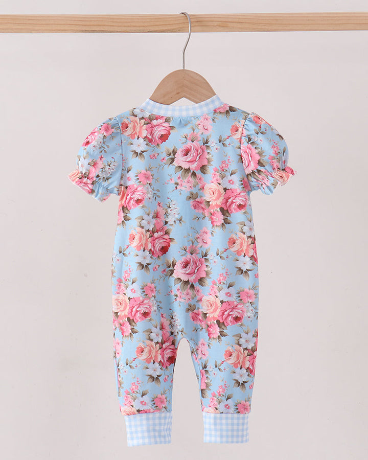 A baby jumpsuit with a light blue background and a pink rose floral print, hanging on a wooden hanger from a wooden clothes rack against a plain light beige wall.