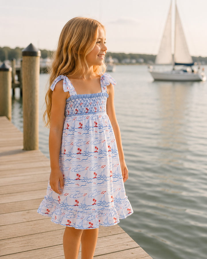 Young girl in a floral dress standing on a dock by a body of water with a sailboat in the background.
