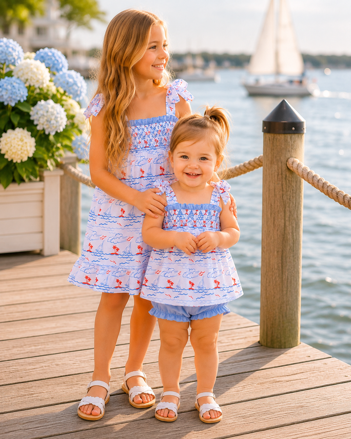Two young girls in matching outfits standing on a dock by water with flowers in the background.