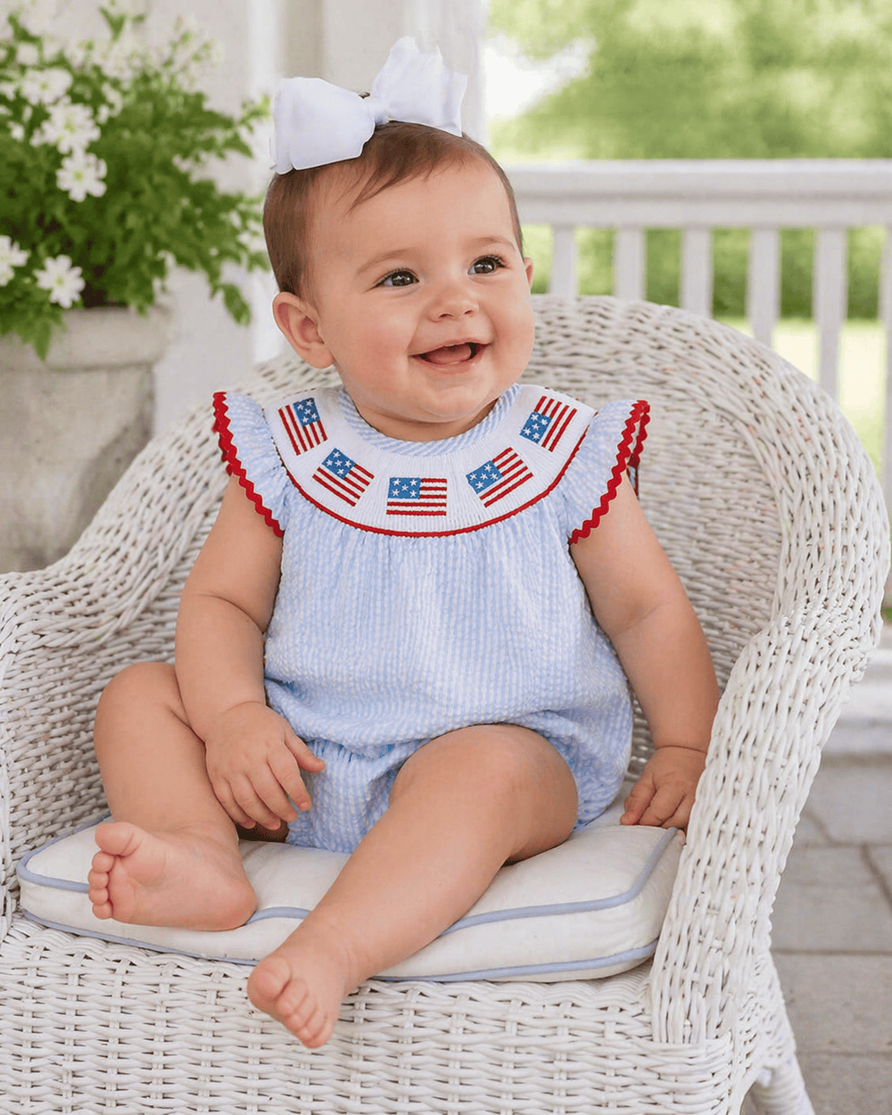 Baby sitting on a wicker chair wearing a blue outfit with American flag details, outdoors.
