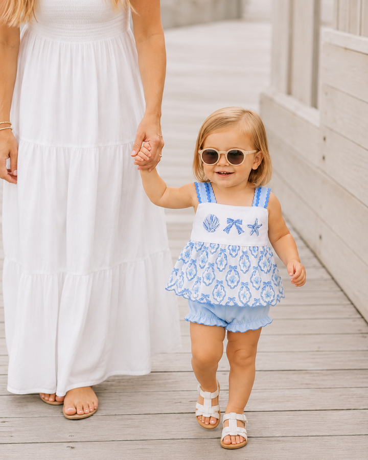 Woman in a white dress holding hands with a young girl in a blue and white outfit on a wooden deck.
