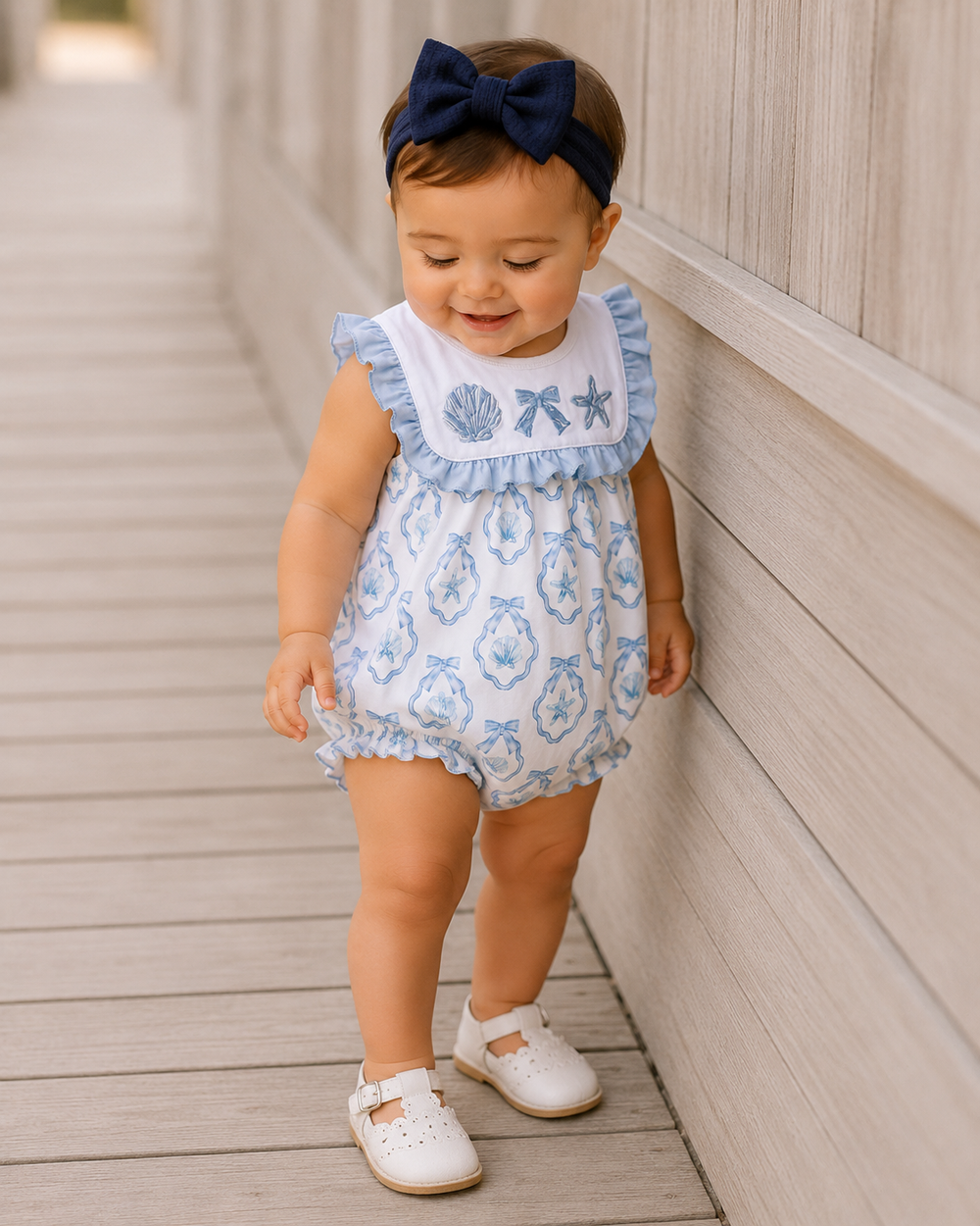 Baby girl in a blue and white romper with a navy bow standing on a wooden deck.