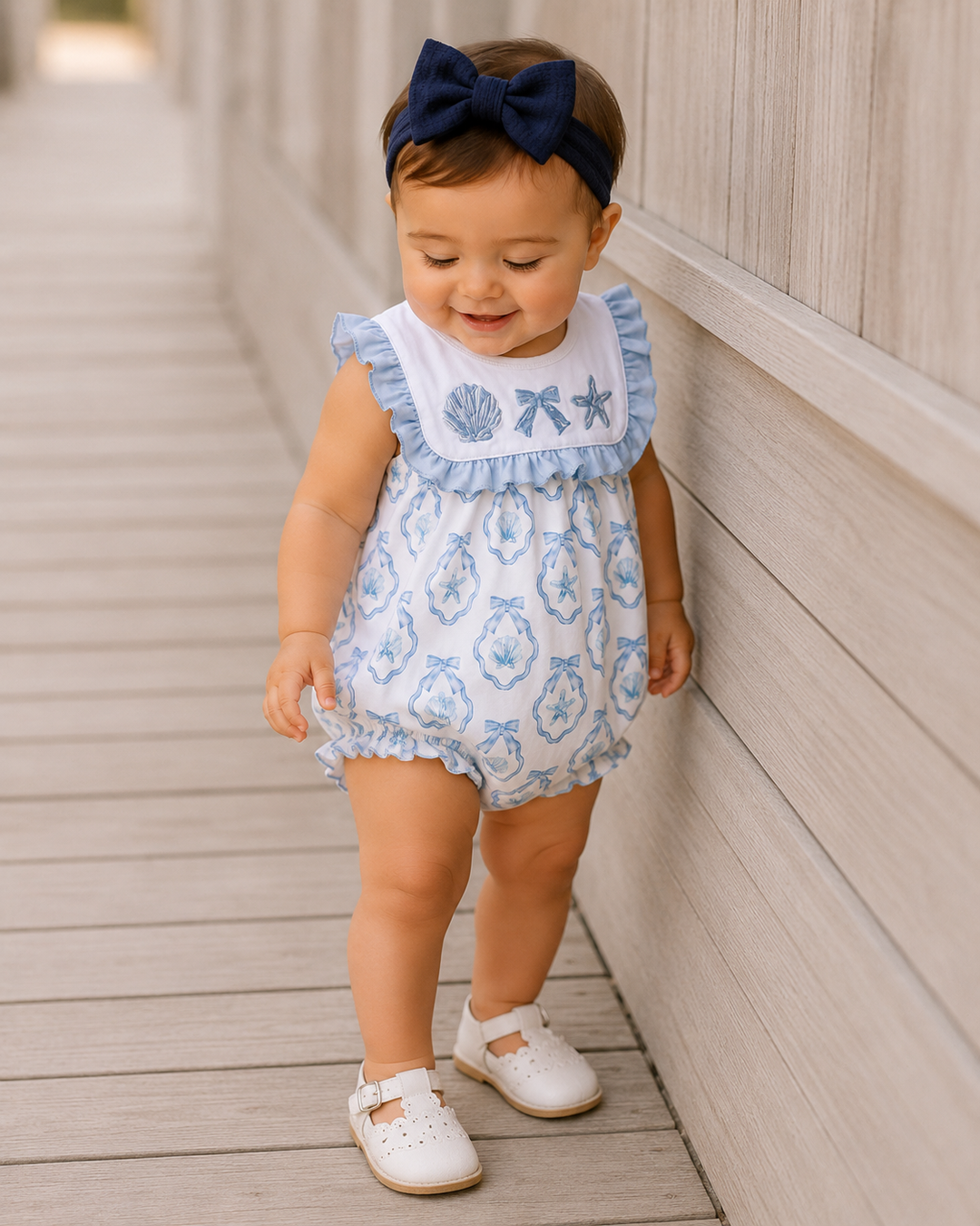 Baby girl in a blue and white romper with a navy bow standing on a wooden deck.