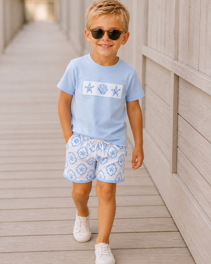 Young boy wearing a light blue t-shirt with a graphic and patterned shorts on a wooden deck.