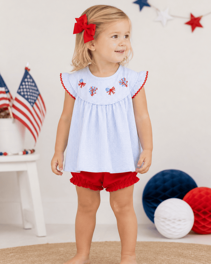 Young girl in a light blue top with red trim and red shorts standing in front of American flags and decorative balloons.
