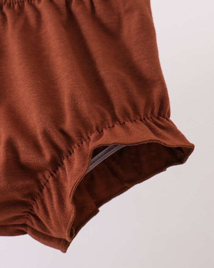 Close-up of a rust-brown fabric garment, showing gathered elastic waistband and ruffled hem detail against a light gray background.