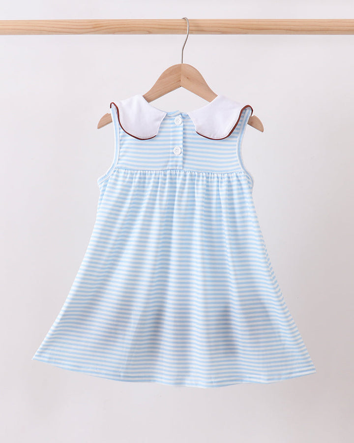A sleeveless dress with light blue and white horizontal stripes, featuring a white Peter Pan collar with red piping, hanging on a wooden hanger against a plain light beige wall.