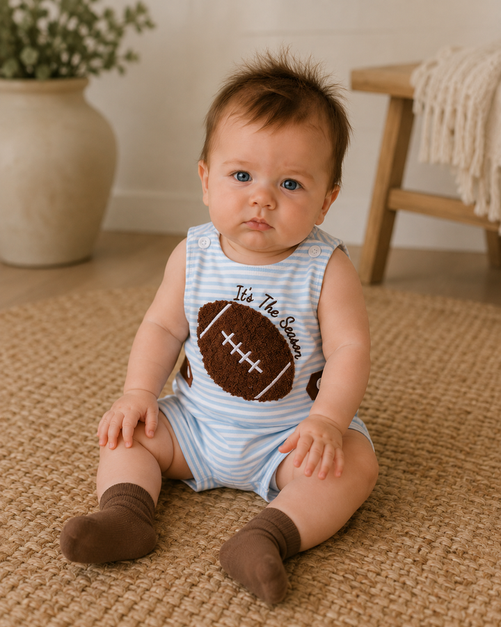 A baby sitting on a woven rug wearing a light blue and white striped sleeveless romper with a brown football graphic and the text &