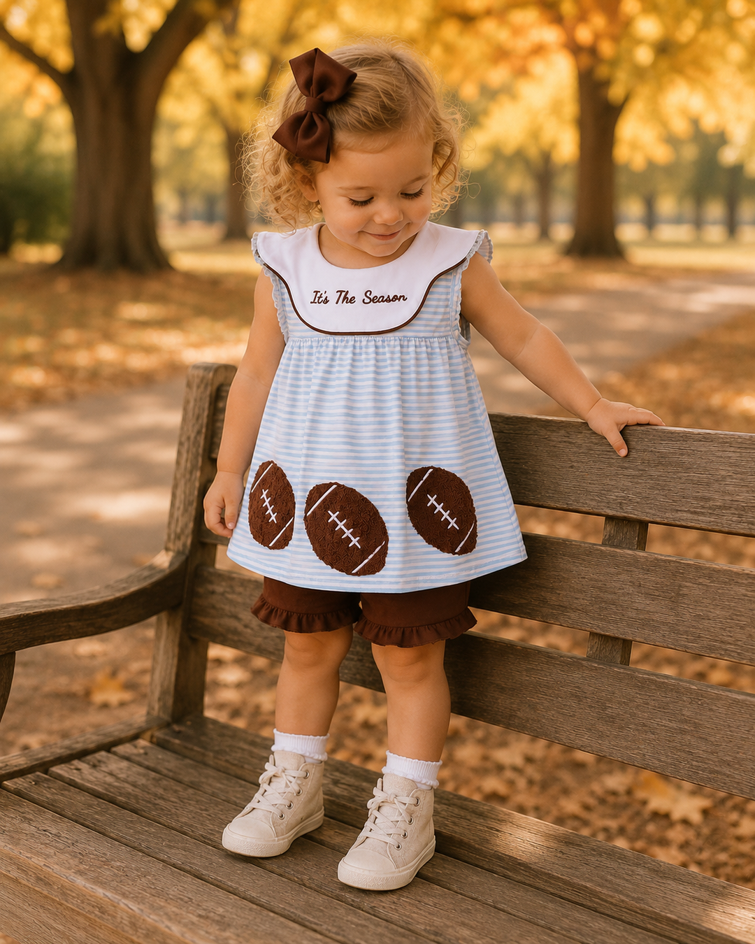 A toddler girl wearing a light blue and white striped dress with brown football appliques and a white bib that says &