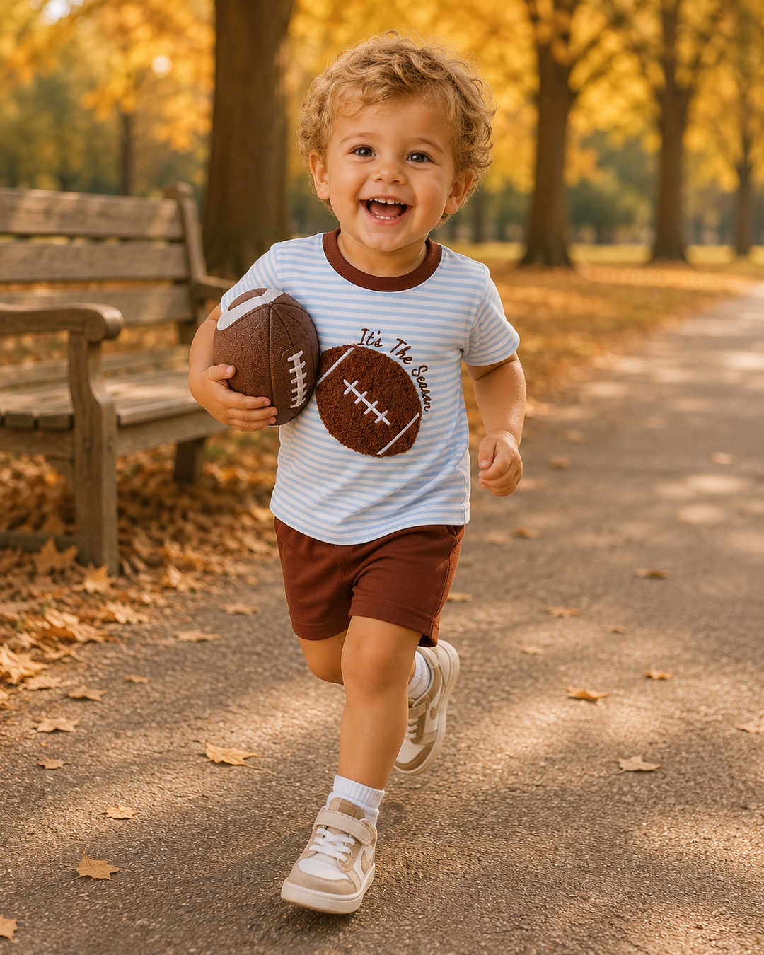 A toddler boy running happily on a tree-lined path in autumn, wearing a white and light blue striped t-shirt with a brown football graphic that reads &