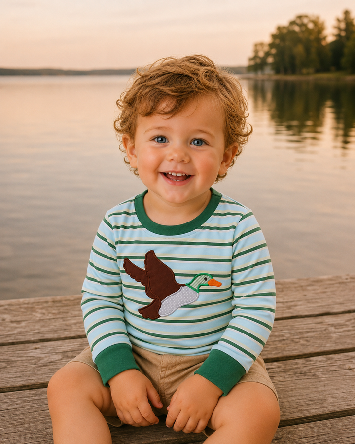 A smiling toddler with curly hair sits on a wooden dock by a lake at sunset, wearing a green and white striped long-sleeve shirt with a flying duck applique and tan shorts.