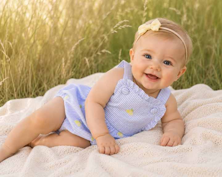 Baby in a blue dress sitting on a blanket in a grassy field