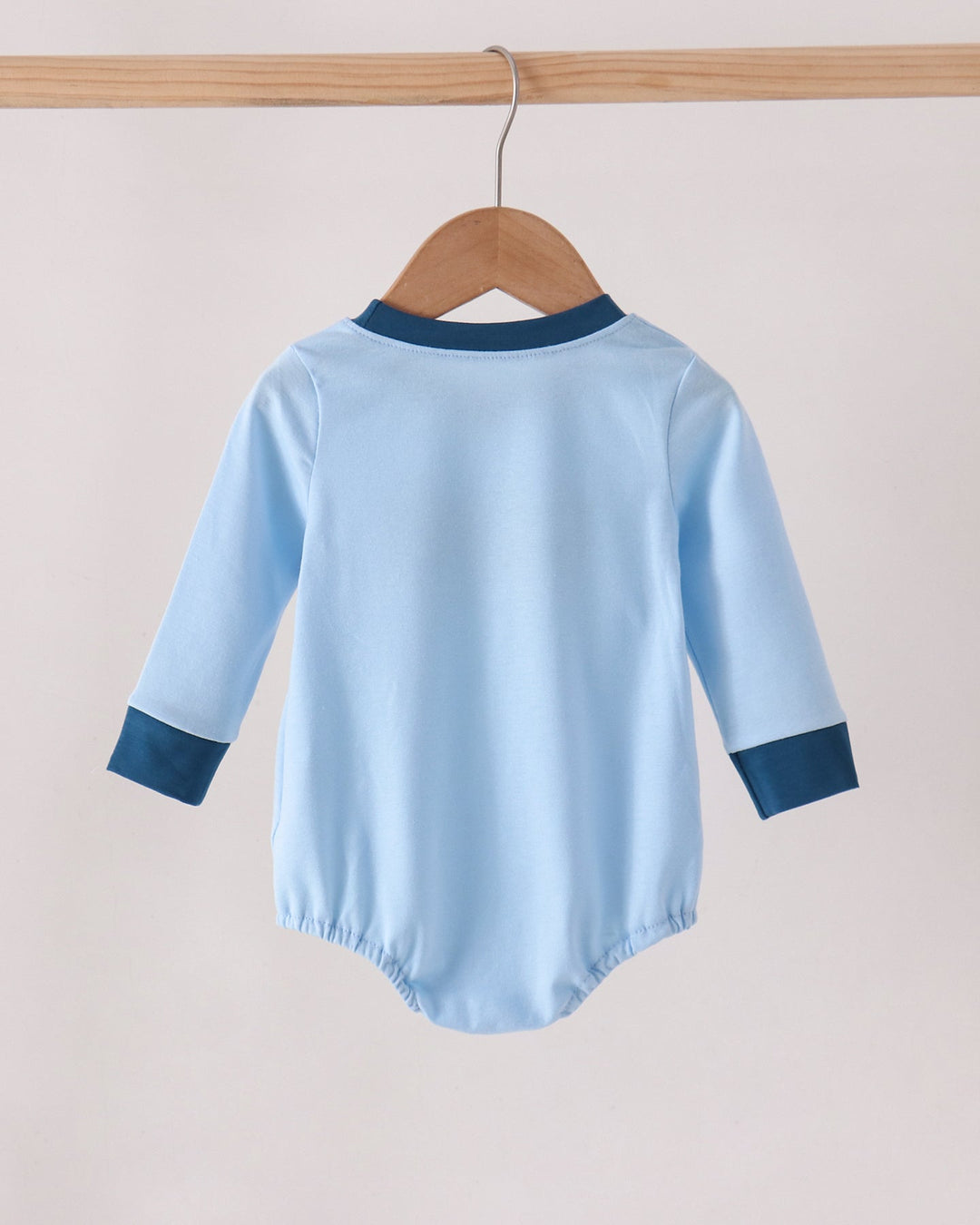 A light blue long-sleeve baby bodysuit shown from the back, hanging on a wooden hanger from a wooden clothes rack against a plain light beige wall.