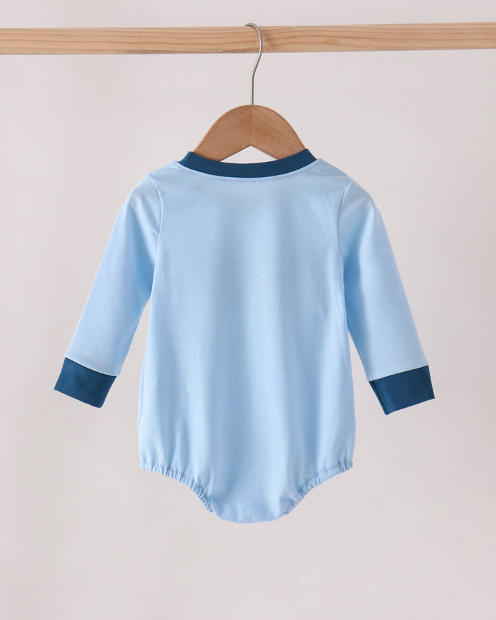 A light blue long-sleeve baby bodysuit shown from the back, hanging on a wooden hanger from a wooden clothes rack against a plain light beige wall.