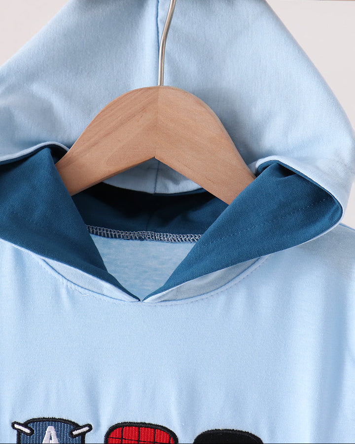 A light blue hooded sweatshirt hanging on a wooden hanger against a plain light background. The hoodie has a contrasting dark blue inner hood lining and a partial graphic print at the bottom showing cartoon-style heads in red, black, and white.