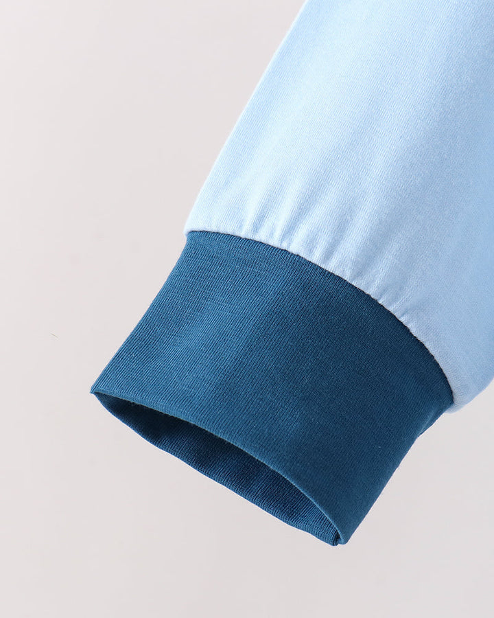 Close-up of a light blue sleeve with a contrasting dark blue ribbed cuff, shown against a plain light beige background.