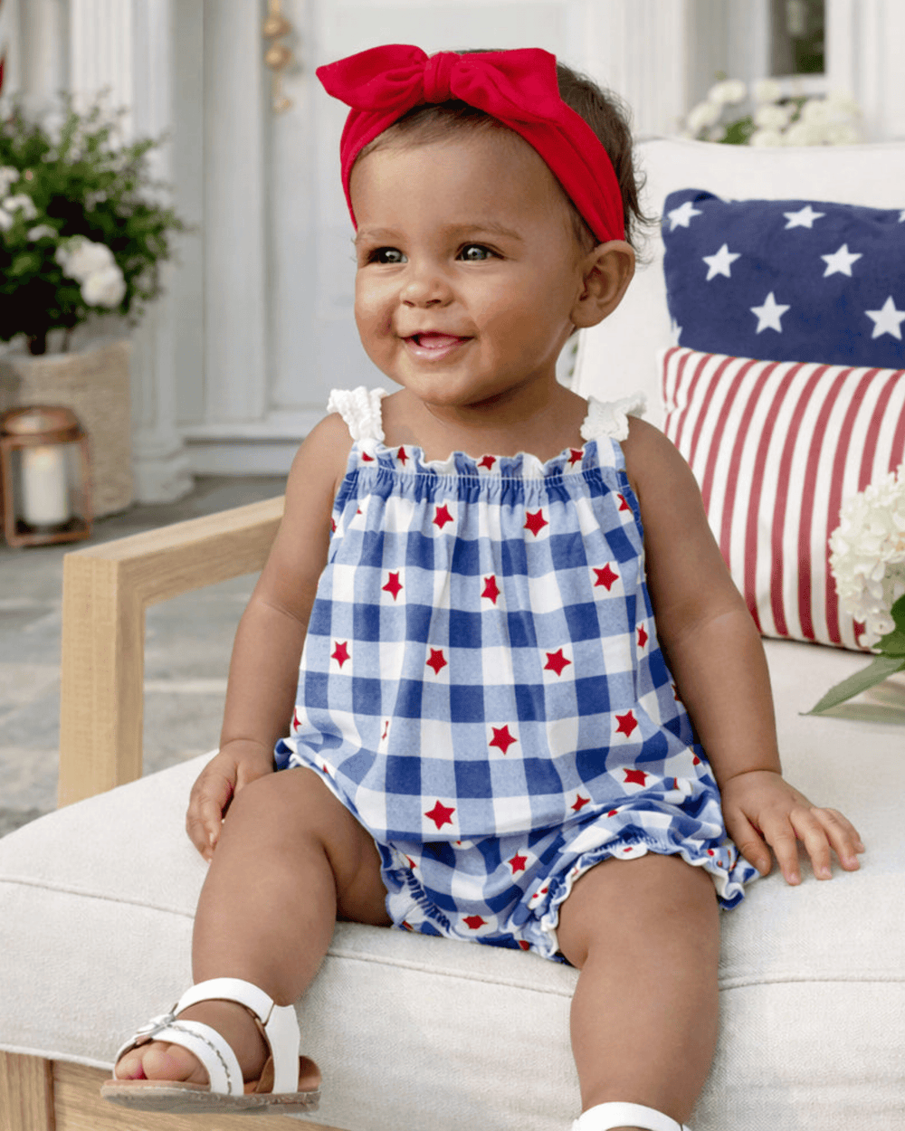 Baby wearing a red, white, and blue checkered romper with star patterns, sitting on a couch.