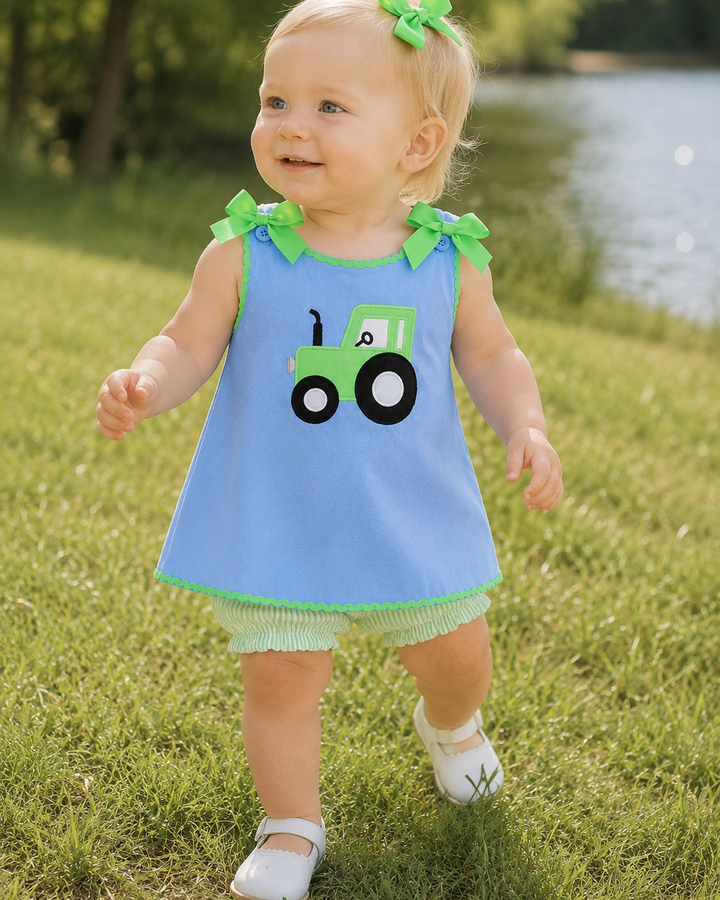 Baby in a blue dress with a green tractor design, standing on grass near a body of water.