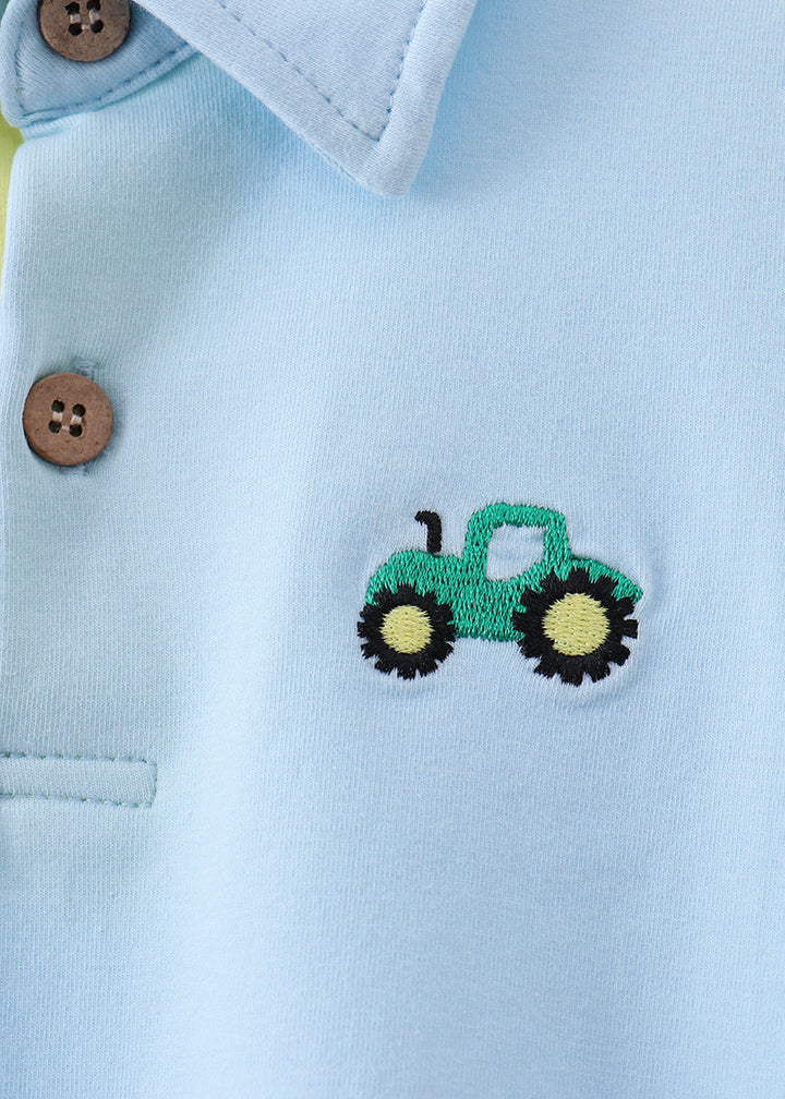 Close-up of a light blue polo shirt featuring an embroidered green tractor with yellow wheels on the chest, with brown buttons on the collar.