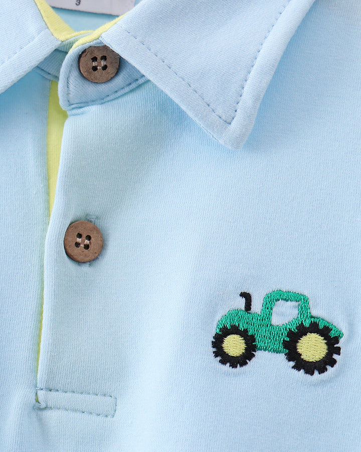 Close-up of a light blue polo shirt with a green embroidered tractor motif on the chest, featuring brown buttons and a yellow inner placket.