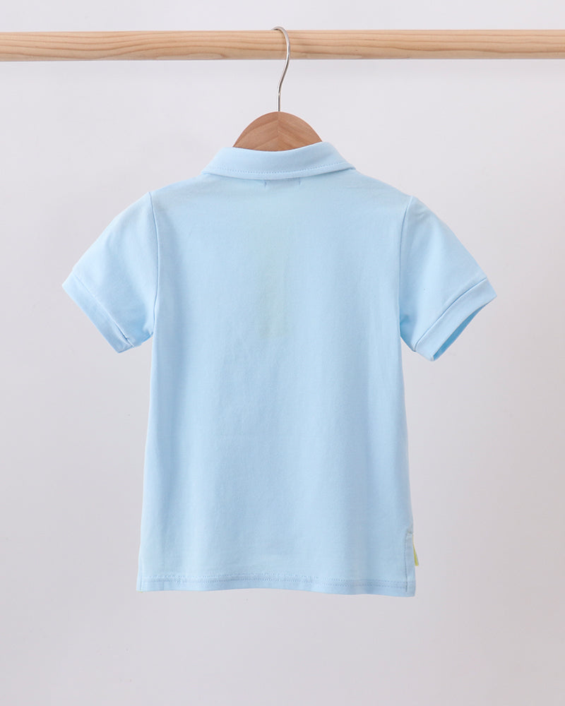 A light blue short-sleeve polo shirt shown from the back, hanging on a wooden hanger from a wooden clothing rack against a plain light gray background.