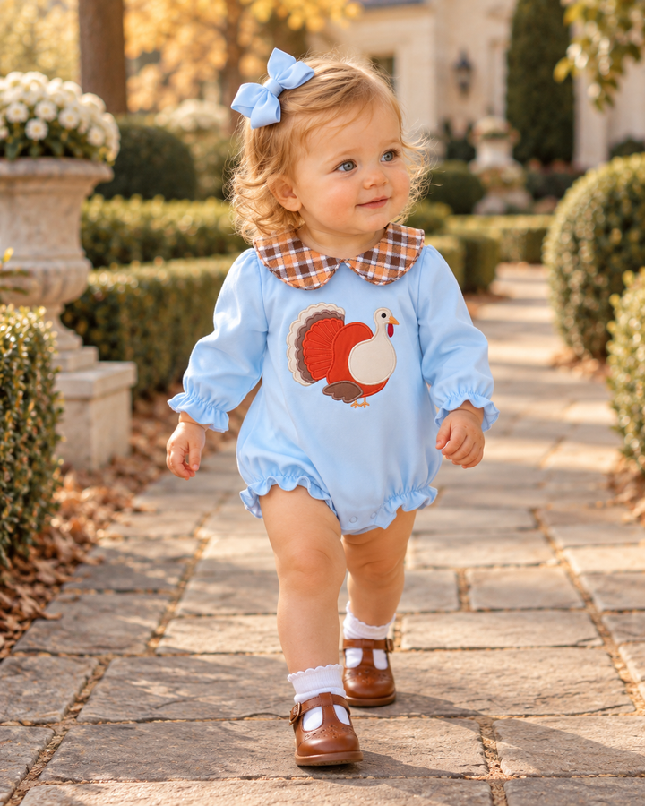Child wearing light blue embroidered turkey romper with brown plaid collar and ruffled legs
