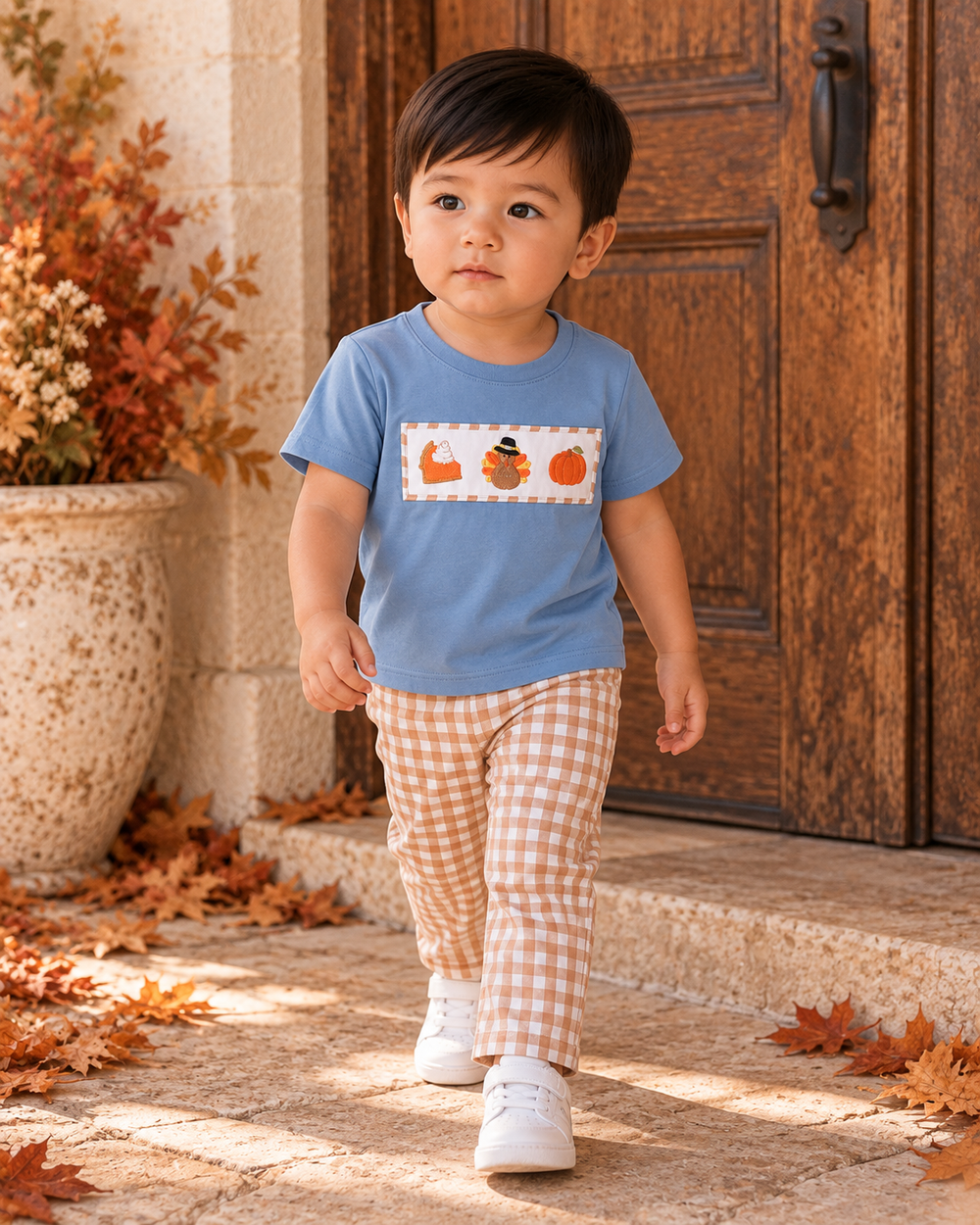 Toddler wearing blue embroidered shirt with turkey and pumpkin graphic and orange gingham pants